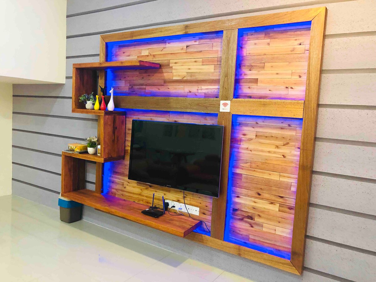 A wooden entertainment unit features a sleek design with a mounted television at its center. LED lighting highlights the wooden panels on either side, while shelves display decorative items and plants, adding a touch of warmth to the living space.