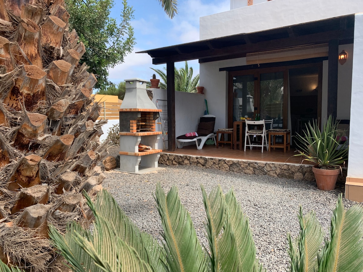 An outdoor terrace is presented with a dining set and a barbecue area, surrounded by palm trees and other greenery. A decorative stone wall is visible, creating a natural barrier. Large windows provide a glimpse of the indoor space beyond.