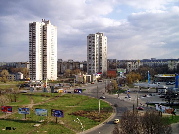 A view showcases tall residential towers flanking a bustling intersection. Surrounding greenery is visible, complemented by multiple billboards and signage. The scene captures an urban environment with various commercial buildings in the background, all under a partly cloudy sky.