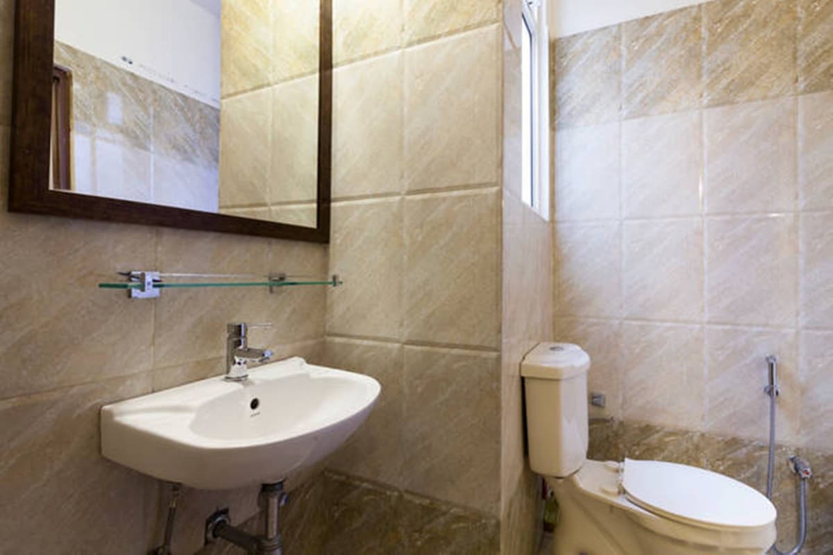 A functional bathroom is depicted, featuring light-toned, textured tiles on the walls. A wall-mounted mirror reflects the space above a compact sink, with a toilet positioned adjacent. A small window allows natural light to enter, illuminating the room.