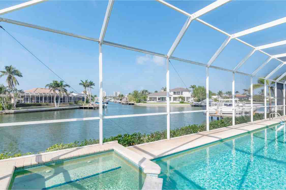 A screened patio area includes a heated pool, providing a view of the canal lined with palm trees. The clear water reflects the vibrant blue sky, while neighboring homes are visible in the distance, highlighting a tranquil waterfront setting.