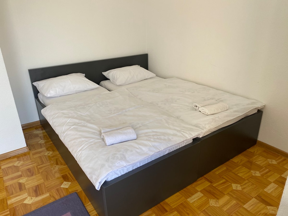 A spacious bed is positioned against a plain wall, featuring fresh white linens and pillows. Two rolled towels rest on the bed, while a wooden floor with a geometric pattern adds a subtle warmth to the room.