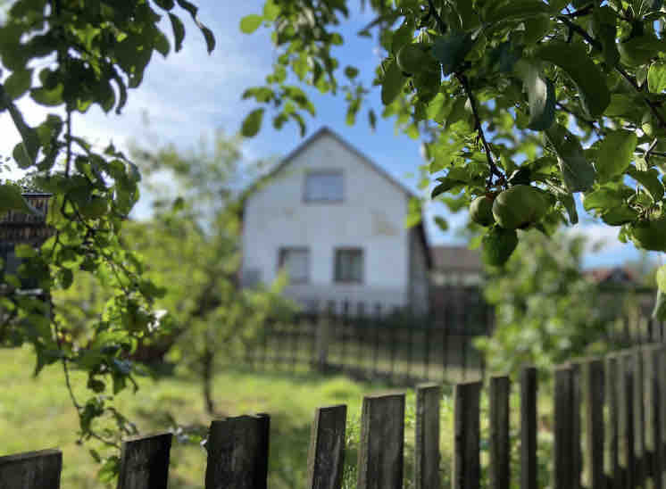 A white cottage is framed by lush green leaves in the foreground. The structure features two windows on the upper floor, with a simple fence surrounding the property and grassy areas visible in the background.
