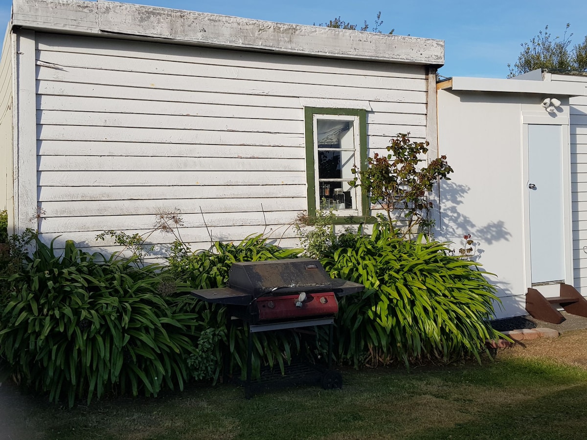 An exterior view of a quaint, weathered structure, framed by lush greenery. A barbecue grill is positioned in front, with a window featuring a green trim visible to the side. The setting presents a relaxed outdoor atmosphere, complemented by a clear blue sky.