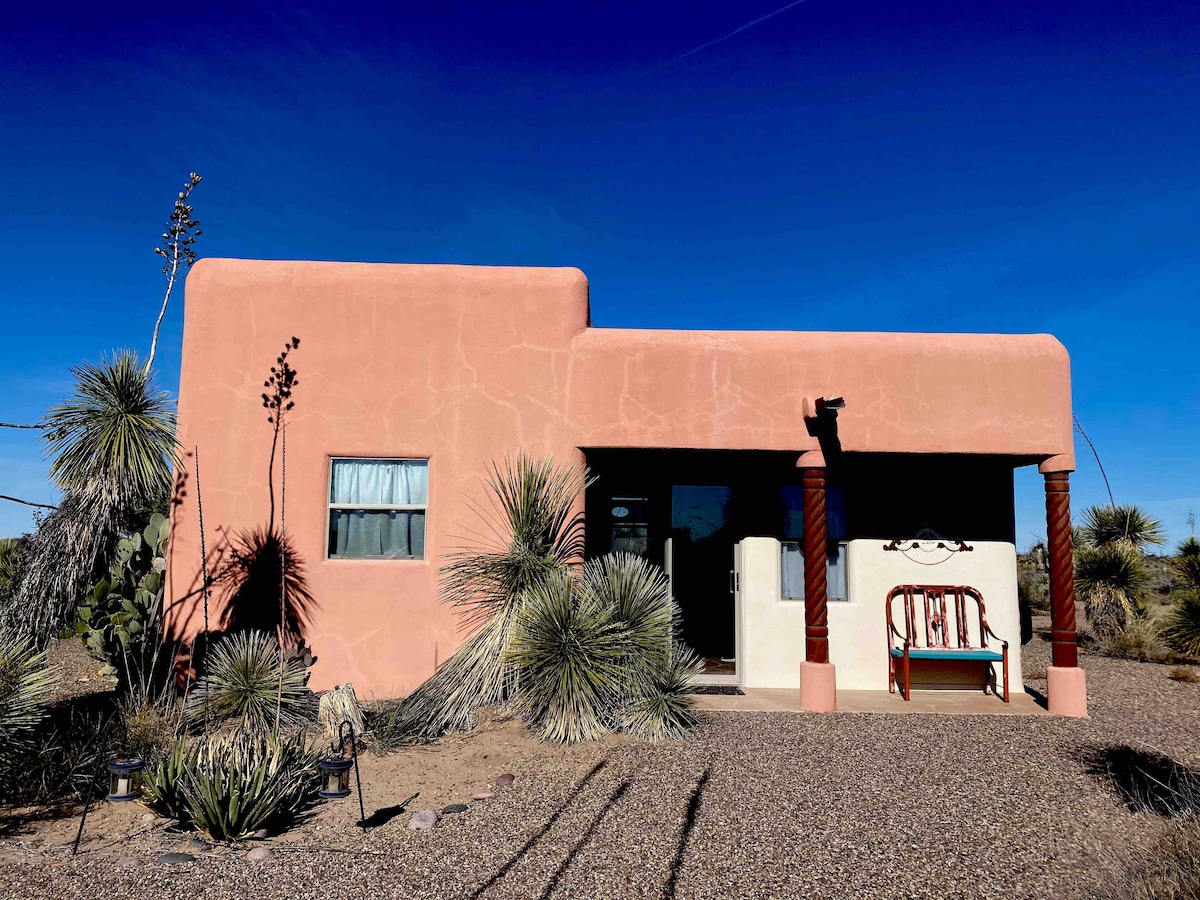 The exterior of a charming casita is presented, featuring a pink adobe finish and traditional wooden columns. Surrounding desert vegetation and a gravel pathway enhance the natural setting. A rustic wooden bench is positioned in front of the entrance, allowing for outdoor relaxation.