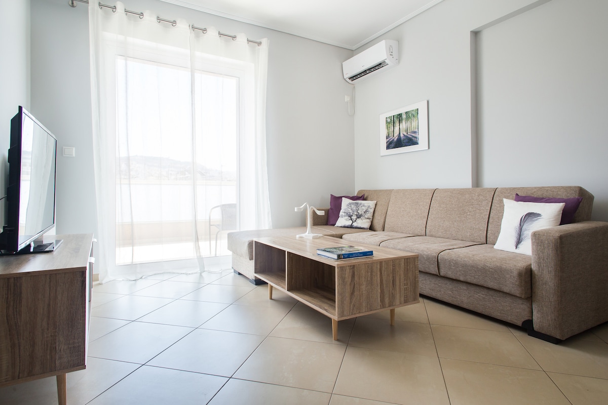 A modern living area is furnished with a spacious sofa and a wooden coffee table. Natural light fills the room through large glass doors, which lead to a balcony. A flat-screen TV is positioned in the corner, with decorative pillows adding a touch of style.
