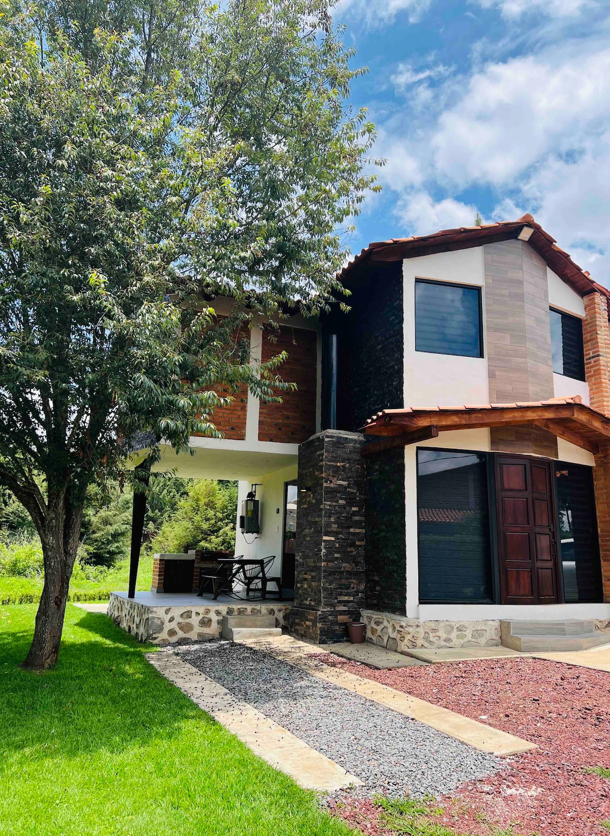 The exterior of the cabin features a blend of stone and wooden elements, complemented by a well-maintained lawn. A pathway made of gravel leads to the entrance, framed by greenery and a prominent tree providing shade. Natural light fills the area under the porch.
