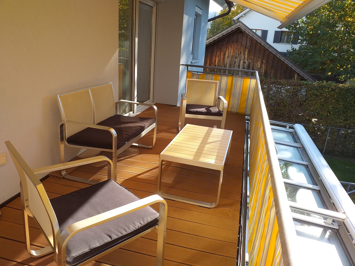 A south-facing terrace features a wooden deck with four metal chairs, each adorned with dark cushions, and a matching coffee table. Bright striped awnings provide shade, while greenery is visible in the background, enhancing the outdoor seating area.