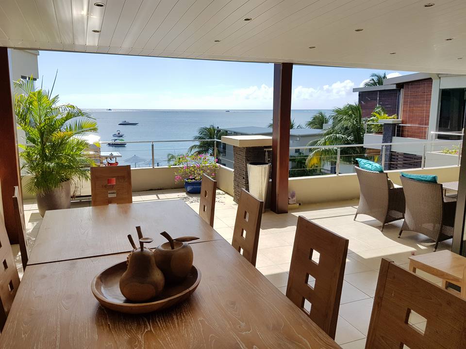 A spacious patio dining area is highlighted by a large wooden table surrounded by chairs. In the background, panoramic sea views are visible, framed by greenery. A decorative bowl containing fruits is placed at the center of the table, adding a touch of warmth to the setting.