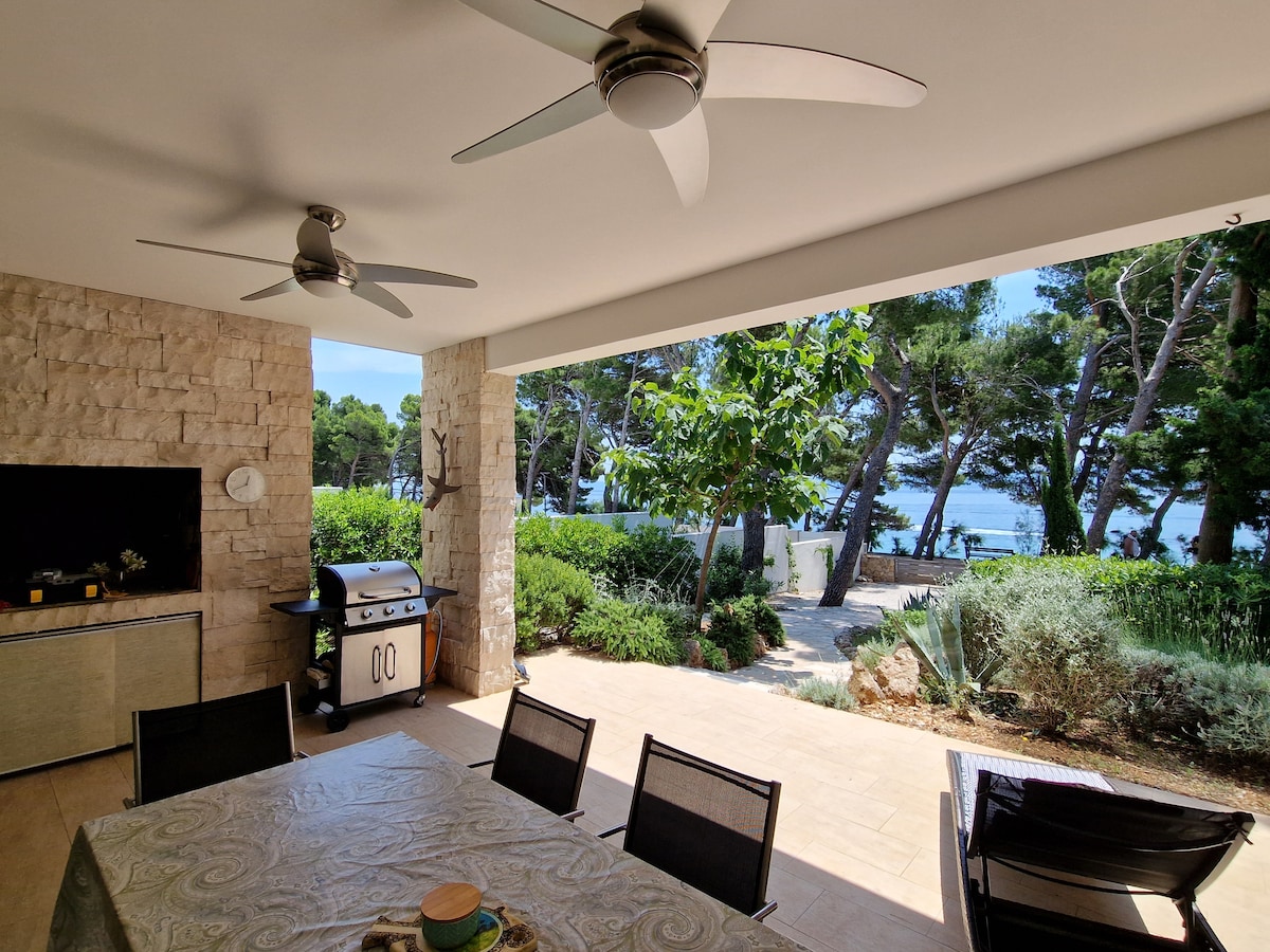 An outdoor dining area features a spacious table surrounded by black chairs. A barbecue grill stands nearby, while lush greenery and trees provide a natural backdrop. The view extends towards a serene water scene, enhancing the inviting outdoor ambiance.