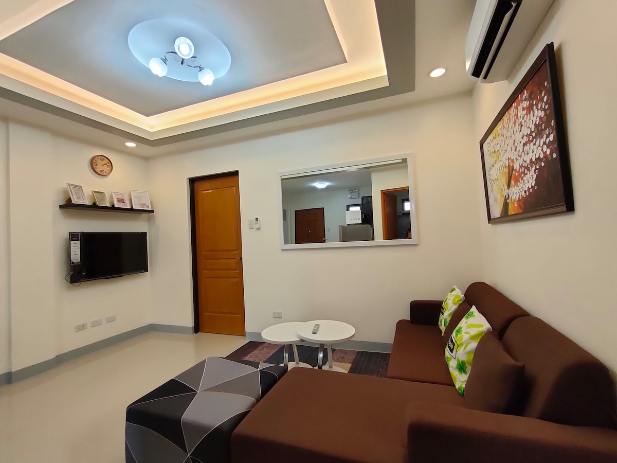 A modern living area is presented with a brown sofa adorned with green-accented cushions. A round white coffee table sits on a textured area rug. A flat-screen TV is mounted on the wall across from the seating, while a mirror reflects the bright, airy ambiance.