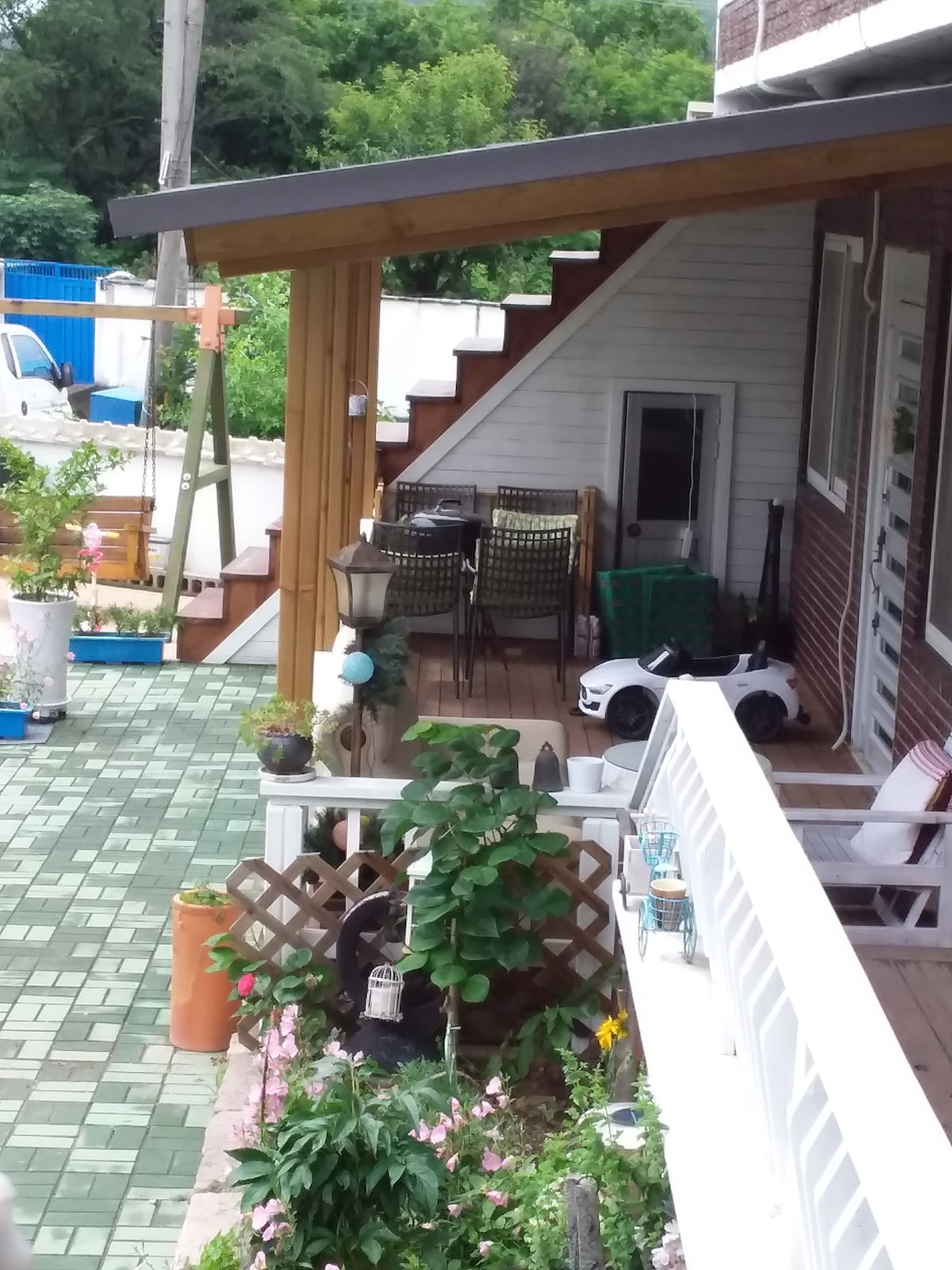 An outdoor area is shown with a wooden deck leading to stairs, surrounded by potted plants and greenery. A small white toy car is parked on the ground, and a dining space with dark chairs is visible in the corner, inviting a sense of relaxation.