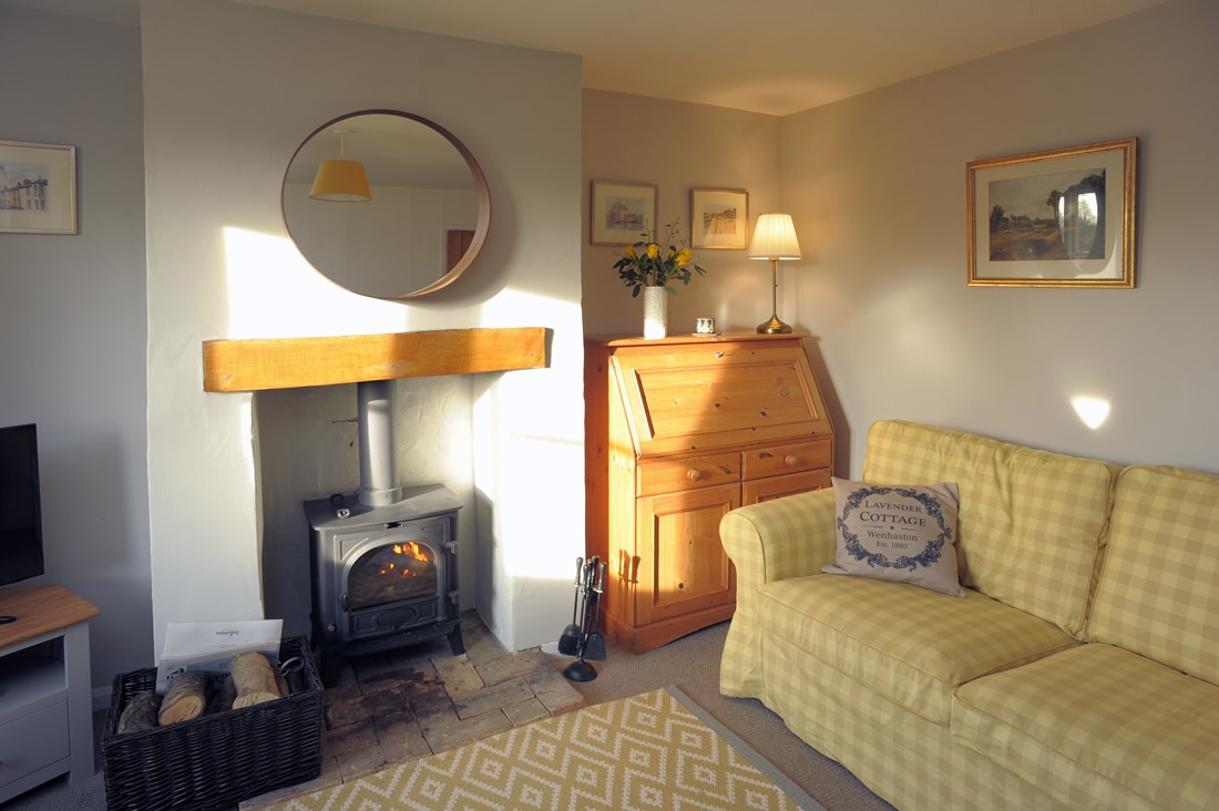 A cozy living room features a log burner, casting a warm glow, and a light yellow checkered sofa inviting relaxation. Soft lighting from a lamp complements the natural light streaming through the window. A wooden sideboard and decorative items add charm to the space.