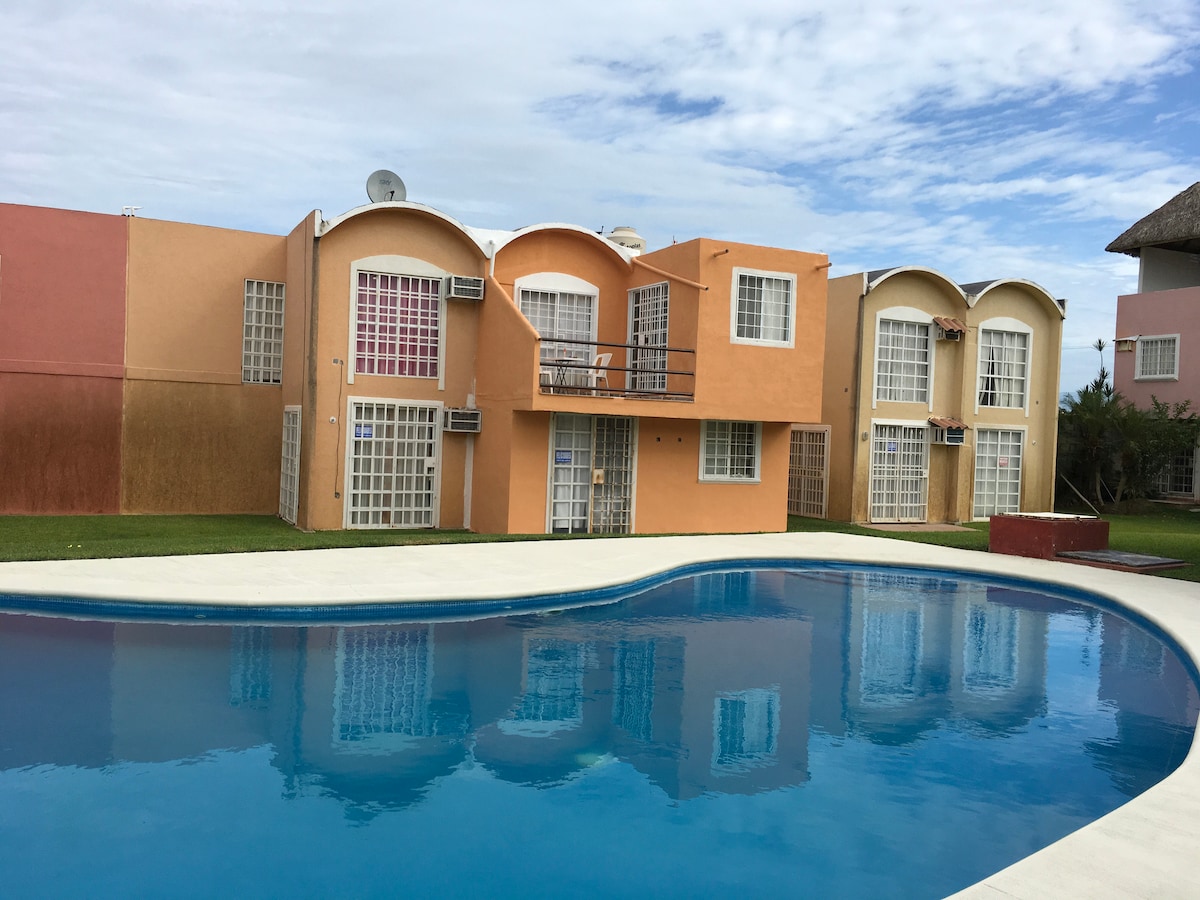 The exterior of a colorful building is reflected in a clear swimming pool. Large, arched windows and air conditioning units are visible on the two-story structure. Lush green grass surrounds the pool, creating a serene environment for relaxation.