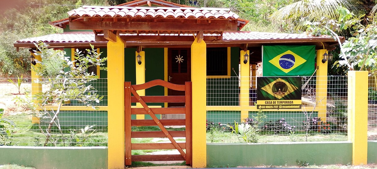 A charming house with a welcoming entrance features a wooden gate and a vibrant Brazilian flag adorning the front. The property is surrounded by a lush green landscape, with yellow and green exterior elements highlighting the home's exterior.