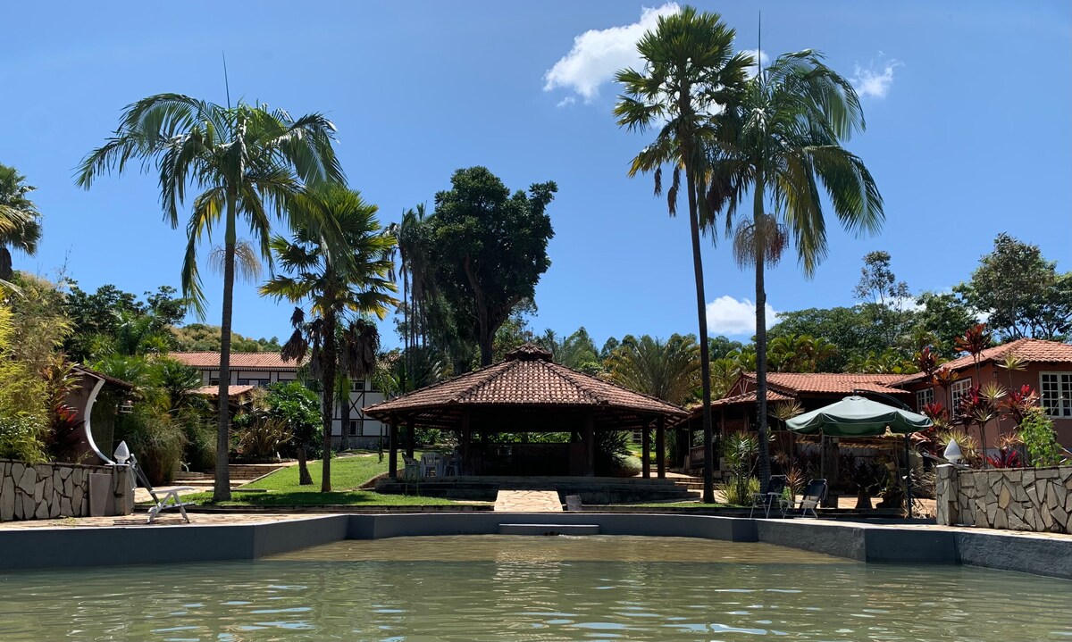 A natural swimming pool is surrounded by tropical palm trees. An open quiosque is visible in the background, framed by lush greenery. The architecture features a traditional roof, complemented by stone pathways leading toward the inviting common areas of the property.