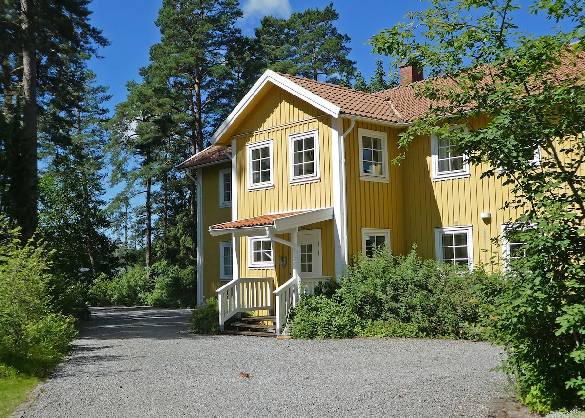 A charming yellow wooden building is surrounded by lush greenery and tall trees. The entryway is highlighted by a welcoming staircase and white railings, inviting guests to explore the natural setting of the property.