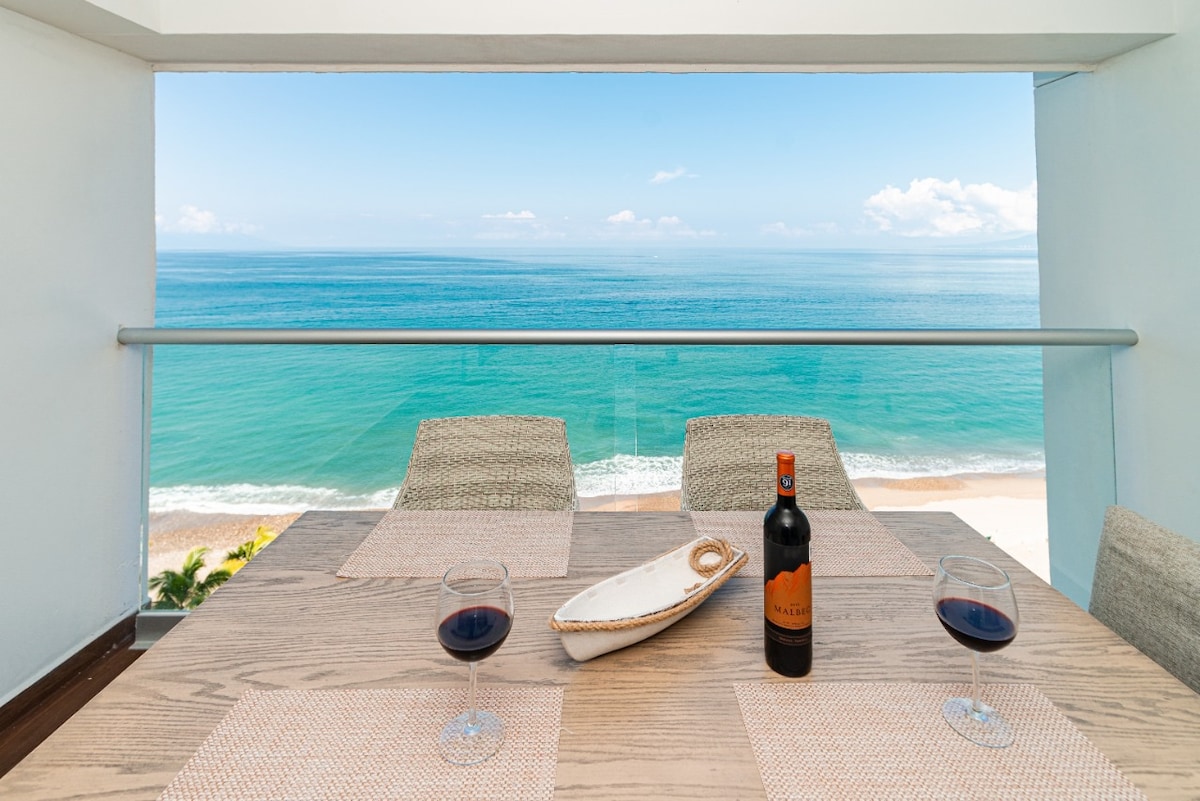 A dining table with two glasses of red wine and a decorative bowl is set on a balcony overlooking the ocean. The expansive view of the sea contrasts with the sandy beach, with clear blue skies above, creating a serene atmosphere.