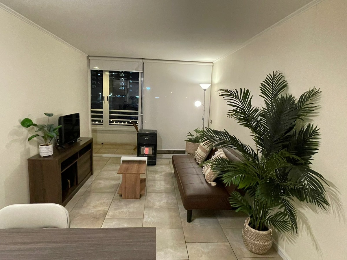 An inviting living area is presented, featuring a comfortable brown sofa paired with decorative cushions. A wooden coffee table is positioned in front. A flat-screen TV is mounted on the wall, complemented by a modern lamp and indoor plants, adding a touch of greenery to the space.
