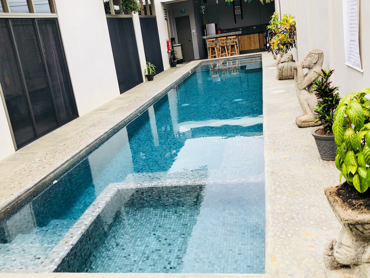 A clear, rectangular swimming pool is bordered by textured concrete and shallow planters filled with green plants. Statues are placed at both ends of the pool. Along one side, a shaded area with seating and a bar is visible, creating a communal space.