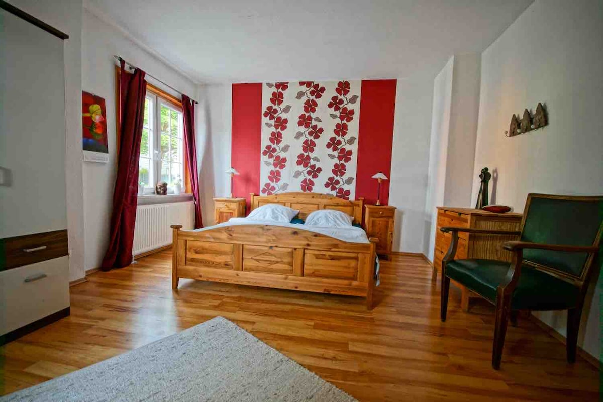 A warm bedroom is presented, featuring a wooden double bed with white bedding. The red flower-patterned accent wall adds color, while wooden furniture, including a nightstand and a chair, complements the decor. Natural light enters through a window, surrounded by red curtains.