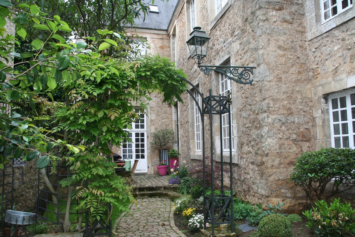 A charming courtyard is surrounded by historic stone walls. Lush greenery frames the entrance, where a decorative lamppost stands. Flowering plants are arranged along the cobblestone pathway, leading to large windows that allow natural light into the nearby interior spaces.