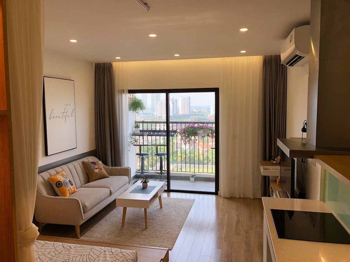 A bright living area features a large window providing city views. A light-colored sofa with decorative pillows is positioned on a light-colored rug. Potted plants are placed on a small table, and curtains frame the window, enhancing the room’s spacious feel.