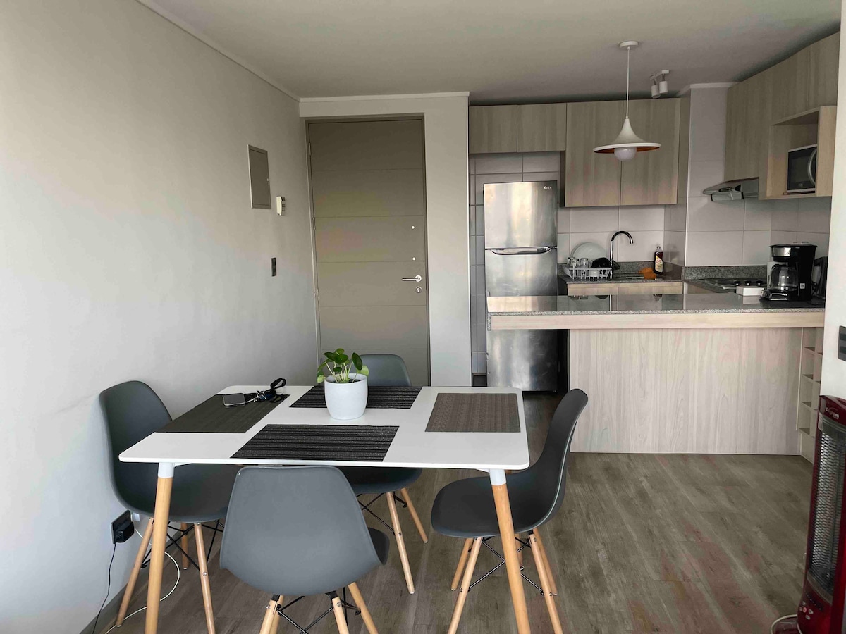 A modern dining area features a white table surrounded by four grey chairs. In the background, a sleek kitchen is visible with a stainless-steel refrigerator and various appliances. Natural light brightens the space through a window, complemented by minimalistic decor.