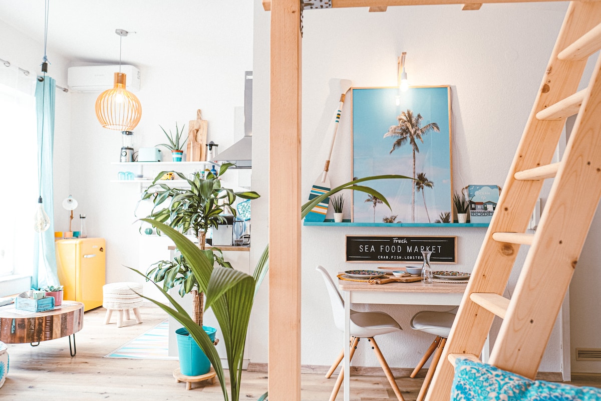 A bright and airy living space features a stylish wooden ladder leading to a loft area. The dining table is set for four and adorned with a tropical-themed artwork. Plants bring a touch of nature, while a vintage yellow refrigerator adds a pop of color in the background.