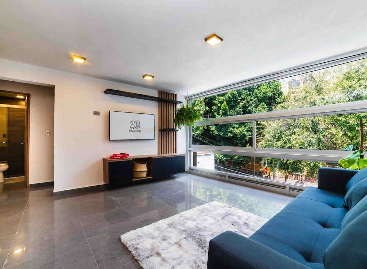 A modern living area features large windows that invite natural light and views of greenery. A plush sofa is positioned across from a wall-mounted television, while a soft area rug lies beneath. The space is complemented by sleek lighting fixtures and minimalistic decor.