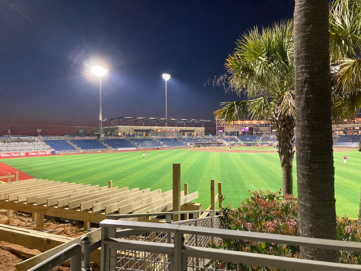 A view of a baseball field lit under night lights, featuring green grass and seating areas in the background. Palm trees are visible in the foreground, adding a tropical touch to the atmosphere. The stadium is filled with a few spectators enjoying the game.