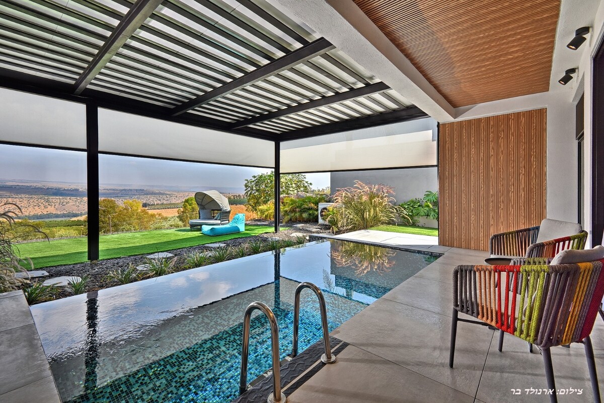 An infinity pool is visible, partially covered by a shaded area, with a tranquil view of natural landscapes in the background. Two wooden chairs with colorful cushions are positioned nearby, along with a lounge chair, surrounded by greenery and modern architectural elements.