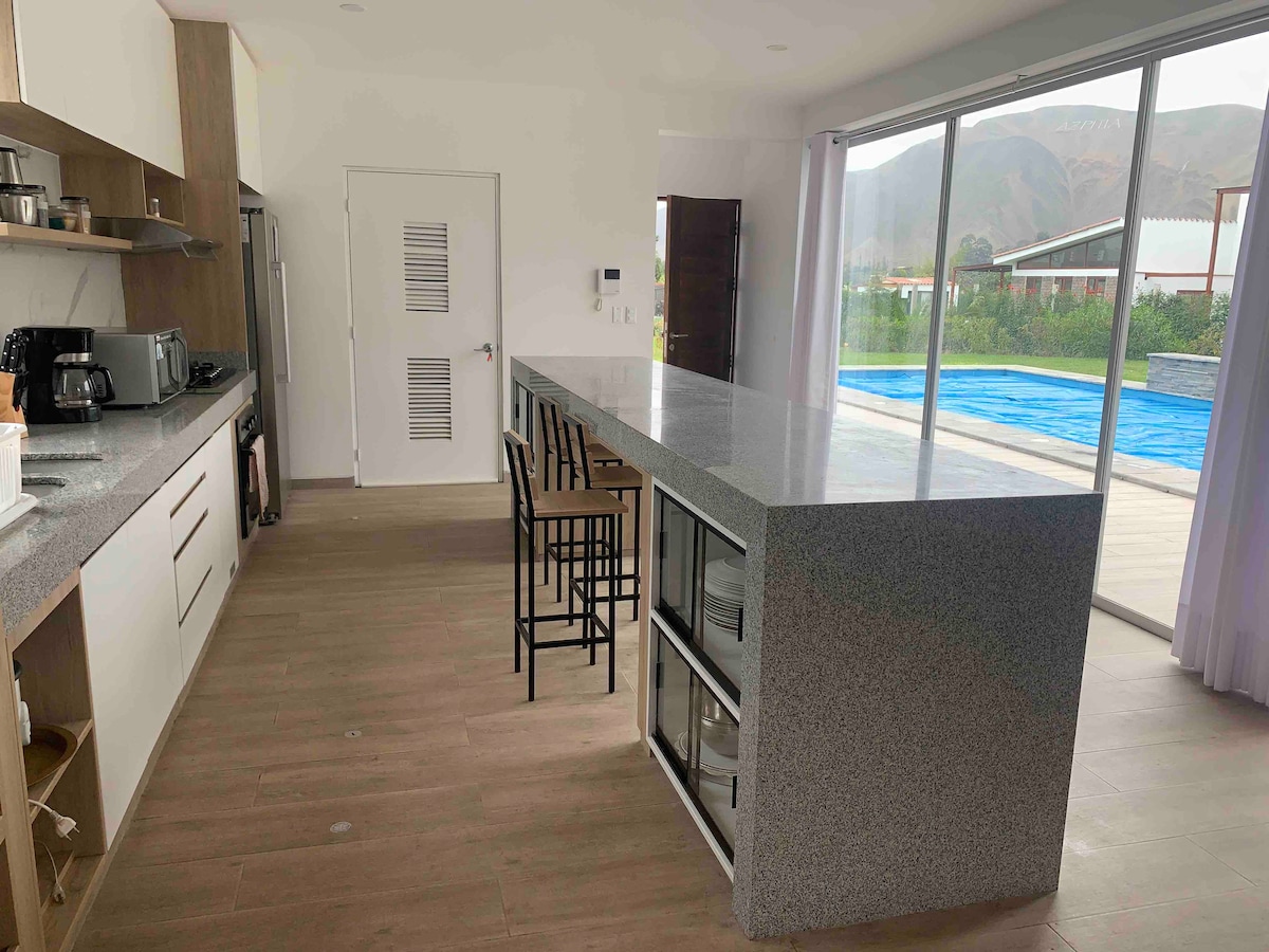 A modern open kitchen features a long granite countertop with bar stools, providing space for casual dining. Appliances such as a coffee maker, toaster, and microwave are visible. A large sliding door allows natural light and views of the outdoor swimming pool area and surrounding hills.