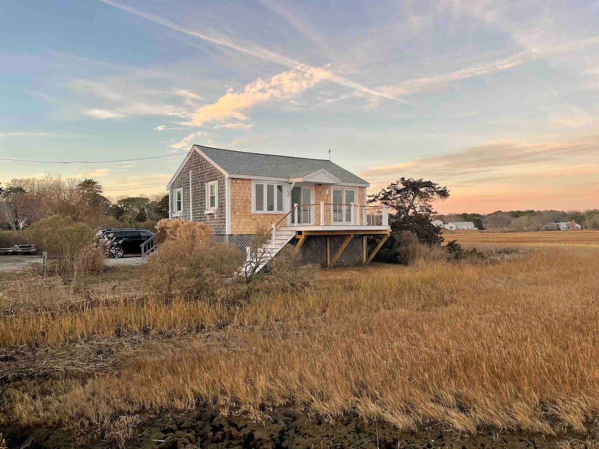 A coastal-style cottage is positioned on stilts, overlooking tall grasses that frame the scenic landscape. A new wooden deck wraps around the front, suggesting a welcoming outdoor space for relaxation. The structure features large windows reflecting the soft colors of sunset.