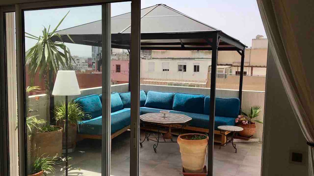 A covered terrace is featured with a large blue seating area surrounded by potted plants. A round table is positioned at the center, offering a space for relaxation. The view includes urban surroundings visible through the glass doors.