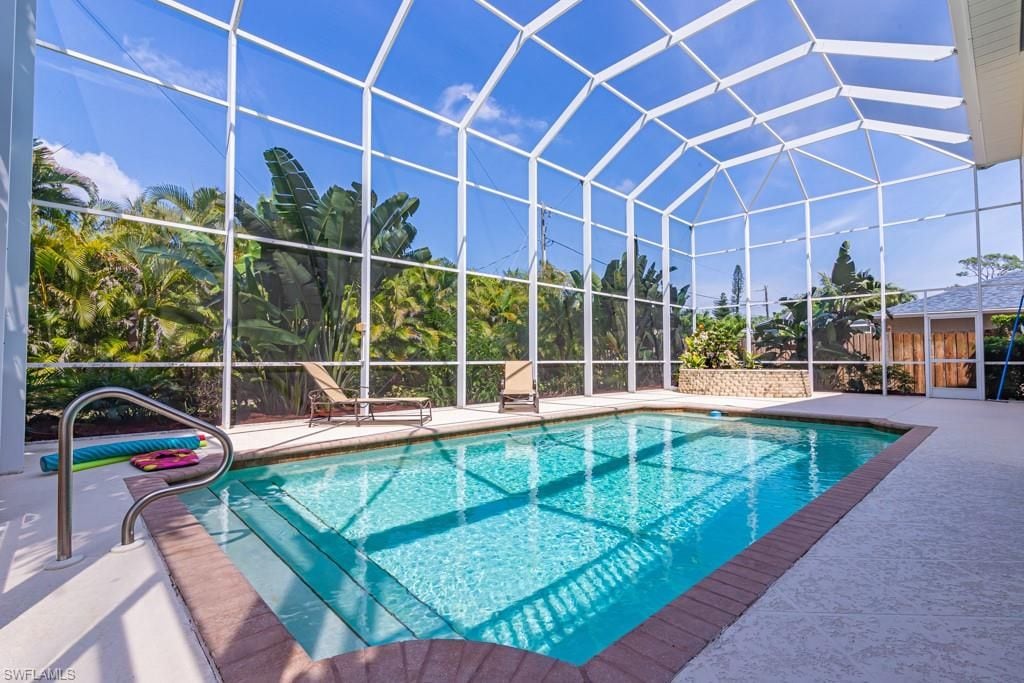 A spacious pool area is surrounded by a protective glass enclosure, with sunlight reflecting off the clear water. Two lounge chairs are positioned beside the pool, and lush greenery can be seen in the background, providing a tranquil setting.
