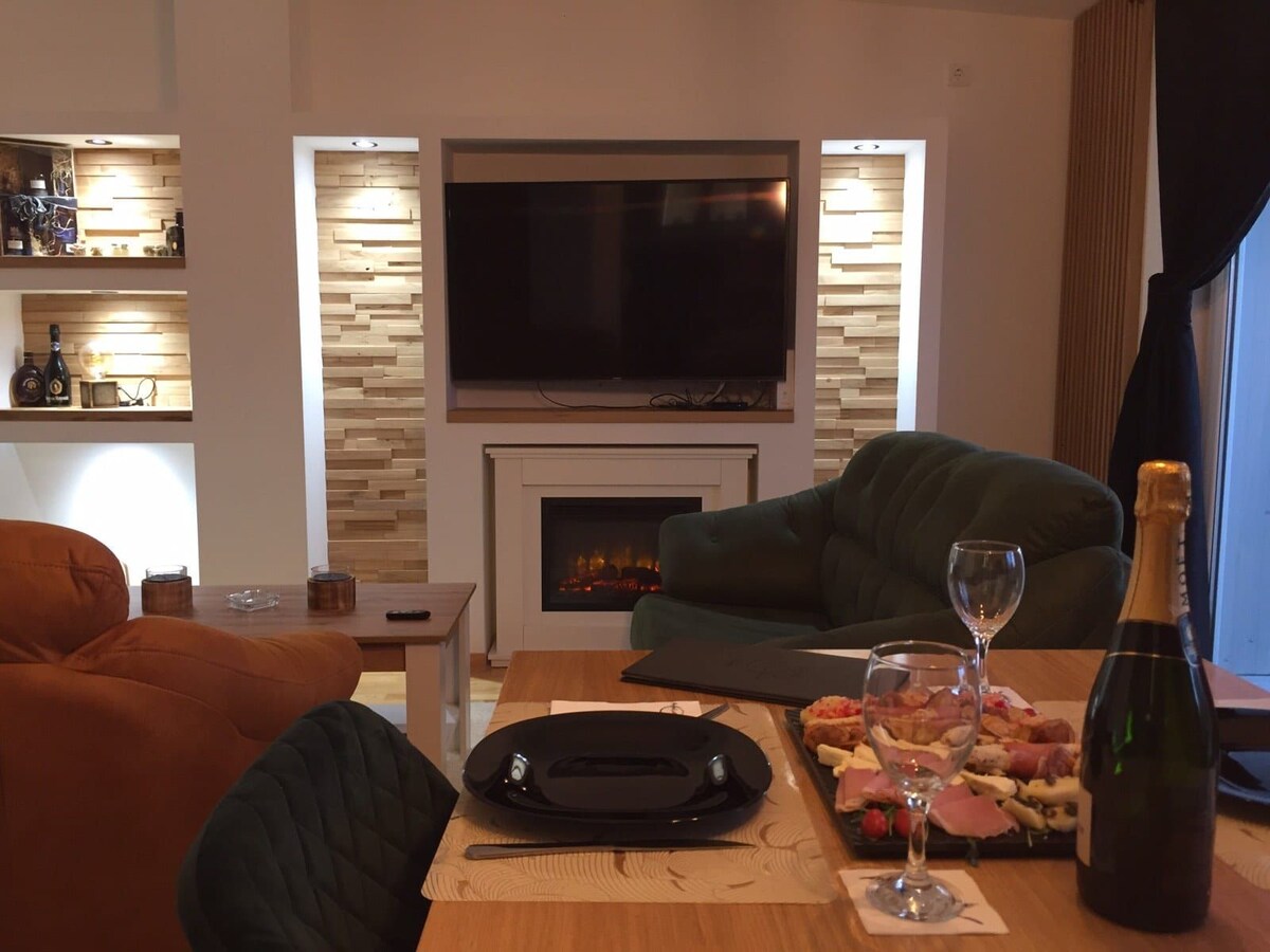 A comfortable living area features a mounted television above a modern fireplace, offering a cozy ambiance. A wooden dining table is set with black plates and elegant glassware, accompanied by a bottle of champagne and an assortment of snacks. Soft seating includes plush sofas in inviting colors.
