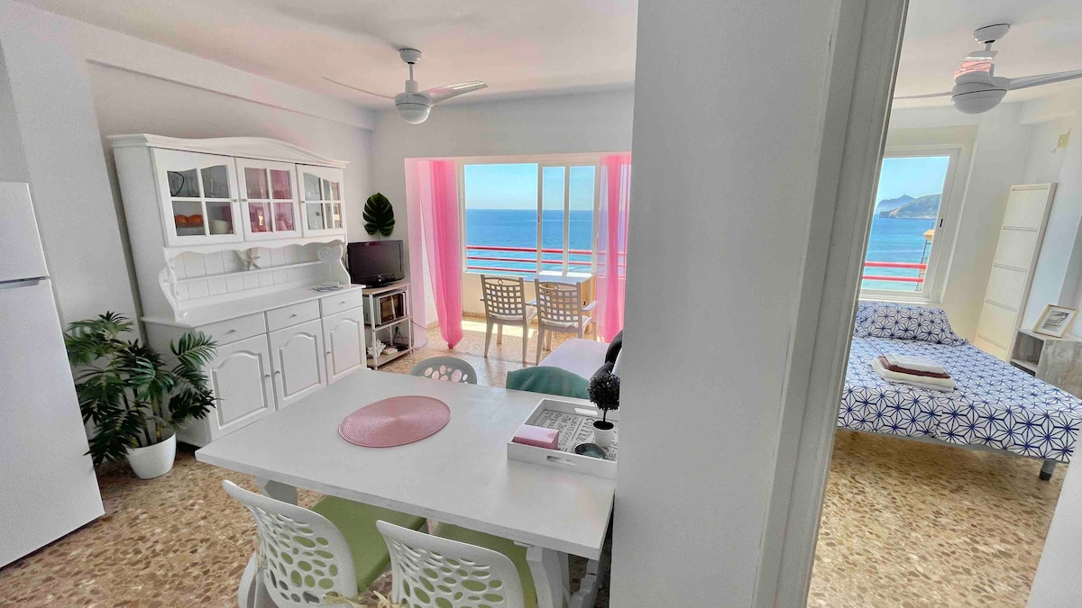 A bright living area features a white dining table surrounded by multicolored chairs. A large window offers panoramic sea views, framed by sheer pink curtains. A comfortable sofa bed is visible, along with a white cabinet and a television, creating a harmonious open space.