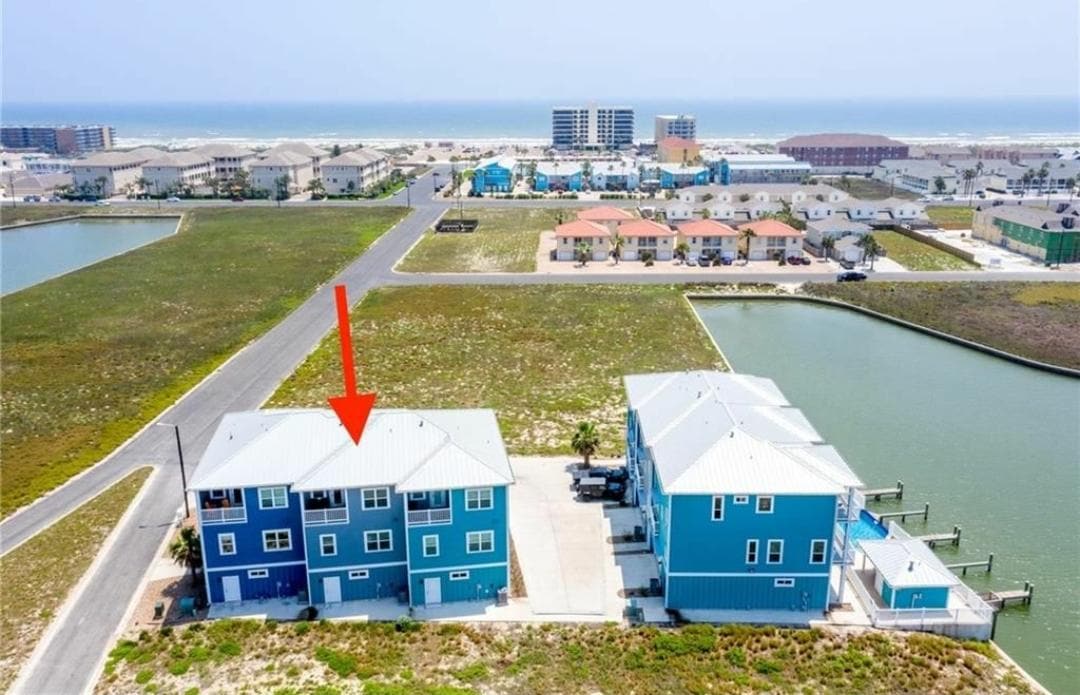 An aerial view showcases a cluster of blue townhomes positioned near a waterway, with the beach visible in the distance. The townhome highlighted by an arrow features a vibrant color and is surrounded by green grass and open spaces.