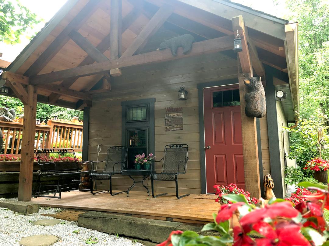 A charming cabin facade is displayed with a wooden overhang and red door. Two black metal chairs are positioned on a wooden deck surrounded by vibrant flowers. Soft gravel paths lead to the entrance, enhancing the inviting atmosphere of the outdoor space.