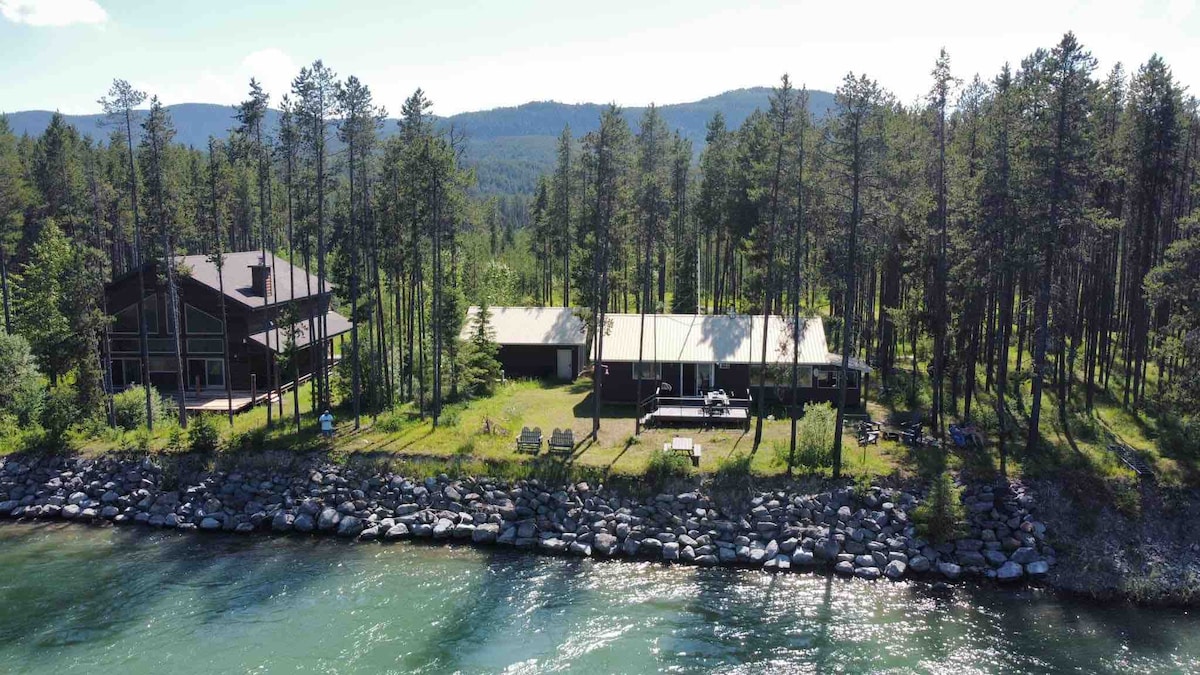 Aerial view of a peaceful retreat nestled among trees along the riverbank. Two separate buildings are visible, complemented by outdoor seating areas on the grassy shore. The clear water reflects the surrounding greenery and distant mountains.