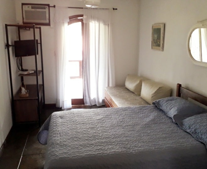 A bright room features a spacious bed covered with a textured blanket. A cozy sofa is positioned near a window adorned with sheer curtains. An air conditioning unit and a wall-mounted TV are visible, providing comfort and entertainment options.