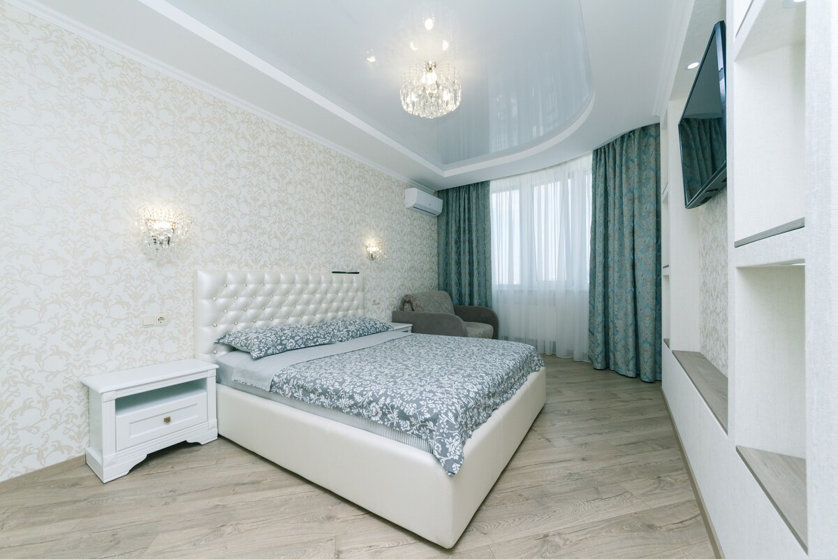 A spacious bedroom features a large, upholstered bed with a decorative bedding set. A chandelier hangs from the ceiling, while a comfortable chair is positioned nearby. Elegant curtains frame the window, allowing natural light to enhance the room's design.