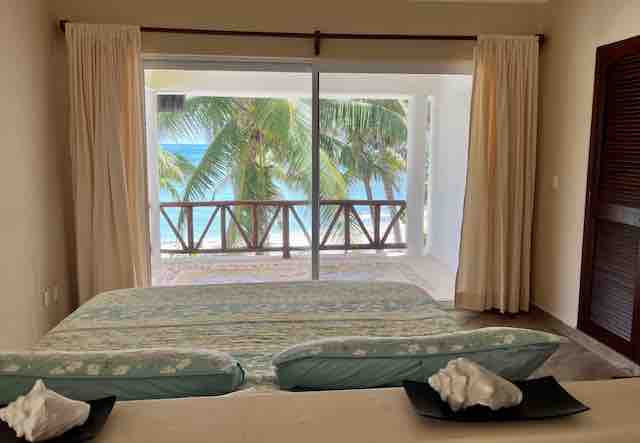 A spacious bedroom features a large bed with light blue bedding. Floor-to-ceiling sliding glass doors reveal a view of palm trees and the ocean. Soft curtains frame the doors, allowing natural light to brighten the space.