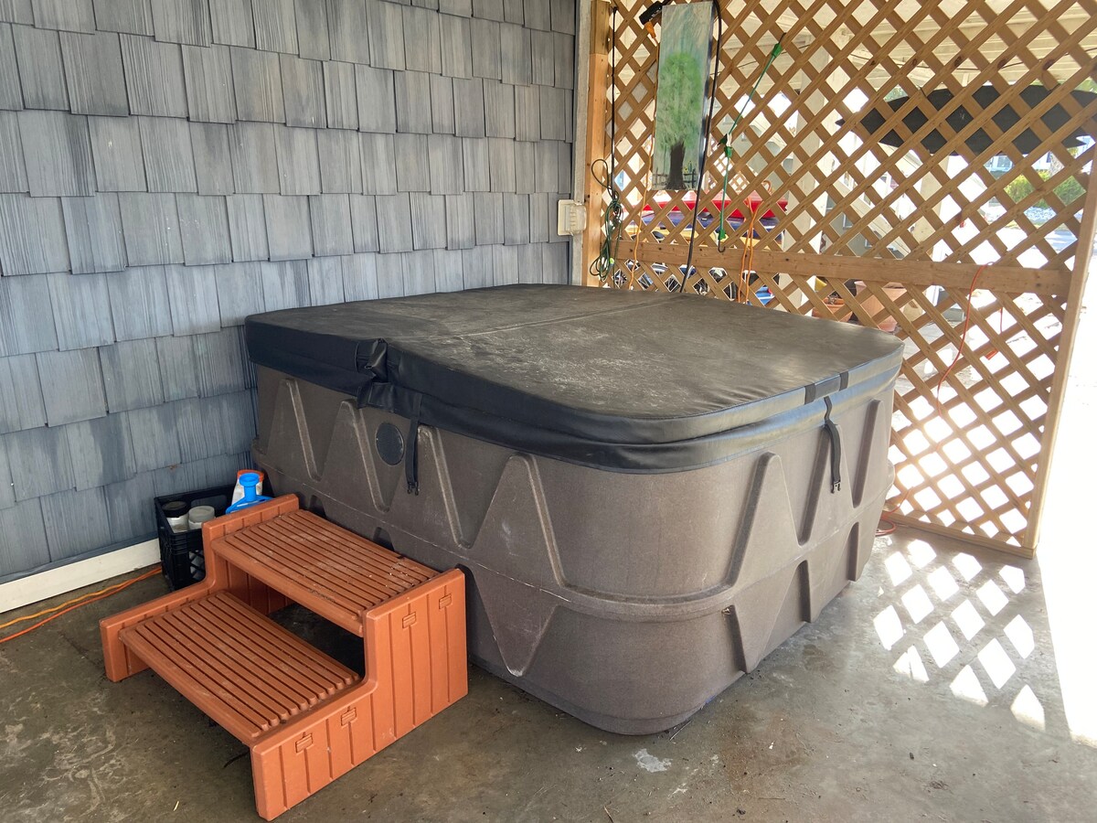 A hot tub is situated on a concrete surface, covered with a black protective lid. Adjacent to the tub, wooden steps lead up to the top. A wooden lattice screen provides some privacy, and items can be seen on the floor nearby.