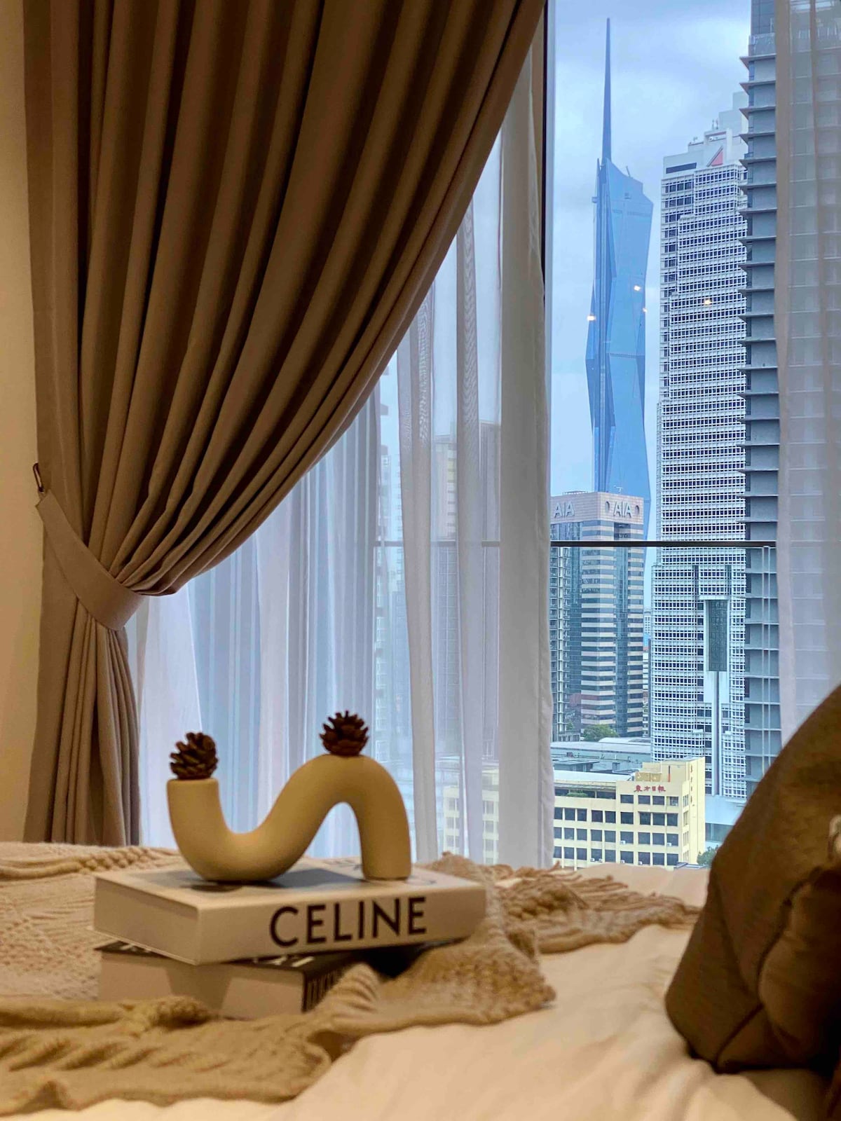 A view from the bedroom features a sculptural decor piece resting on a stack of books. Floor-to-ceiling curtains frame a stunning cityscape, showcasing modern skyscrapers, including the recognizable Petronas Twin Towers in the distance.