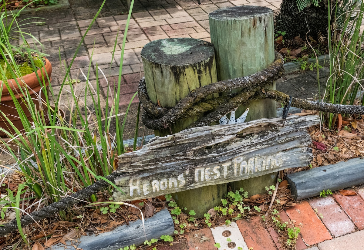 A rustic wooden sign reading 'Herons Nest' is displayed on a weathered log, supported by two cylindrical wooden posts. Surrounding greenery and textured outdoor flooring enhance the natural ambiance of the entrance to the condo.