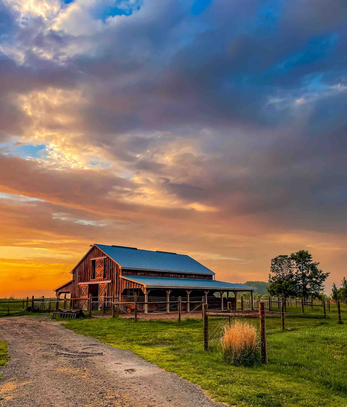 A rustic barn stands against a vibrant sunset sky, showcasing an array of warm colors and clouds. The structure features a wood façade with a sloped roof. A dirt path leads toward the barn, bordered by green grass and a wooden fence.