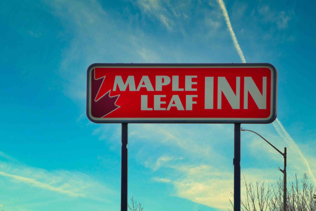 A prominent roadside sign displays the name 'Maple Leaf Inn' in bold letters. The sign features a red background with white text, complemented by a stylized maple leaf graphic. A clear blue sky with light clouds serves as the backdrop.