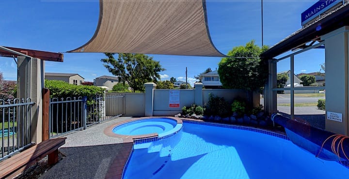 An outdoor area features a shared pool and hot tub, surrounded by a blend of concrete and stone landscaping. Shade sails provide relief from the sun, while trees and a fence frame the space. A sign indicates pool rules, fostering a welcoming environment.